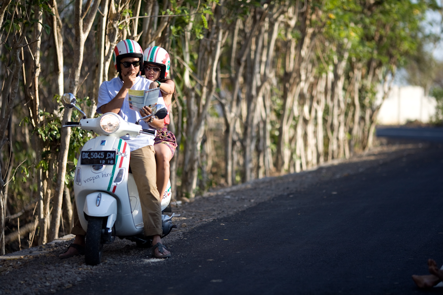 Pasangan mengenakan helm warna-warni sedang melihat peta di atas Vespa putih di jalan pedesaan Jakarta dalam sesi foto prewedding oleh MOTRETKU.
